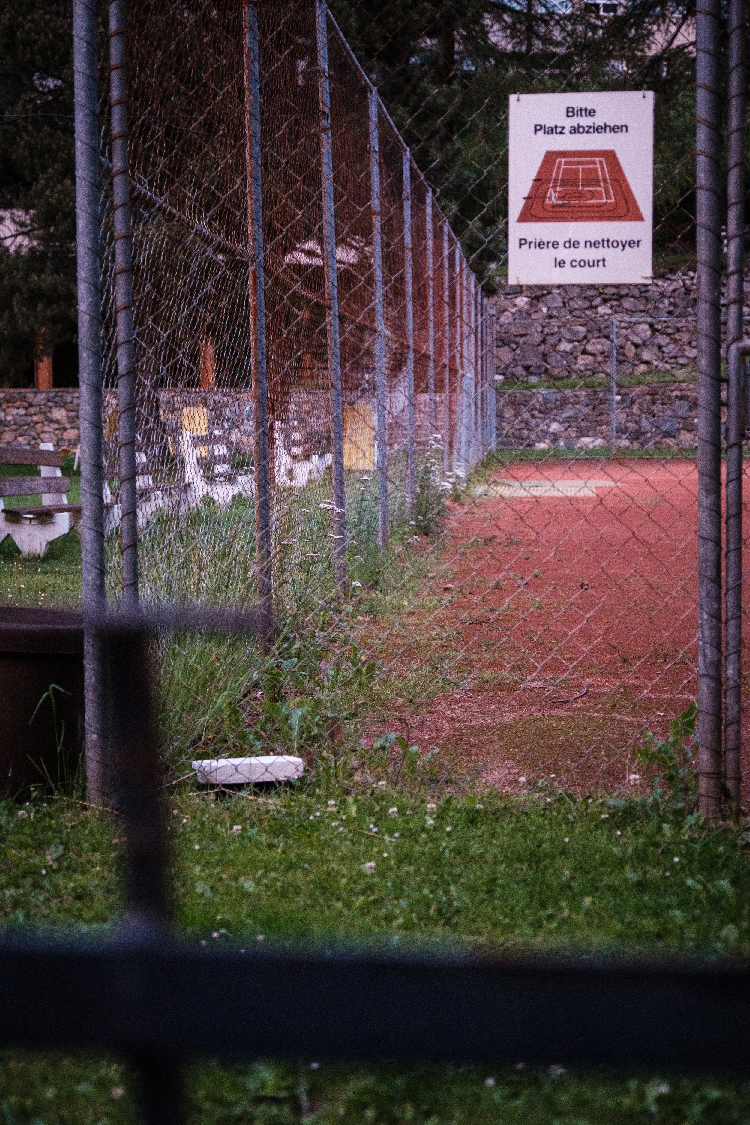 Ein umzäuntes Tennisfeld mit einem Warnschild, das auf Deutsch und Französisch anzeigt, den Platz zu räumen und den Platz zu reinigen.