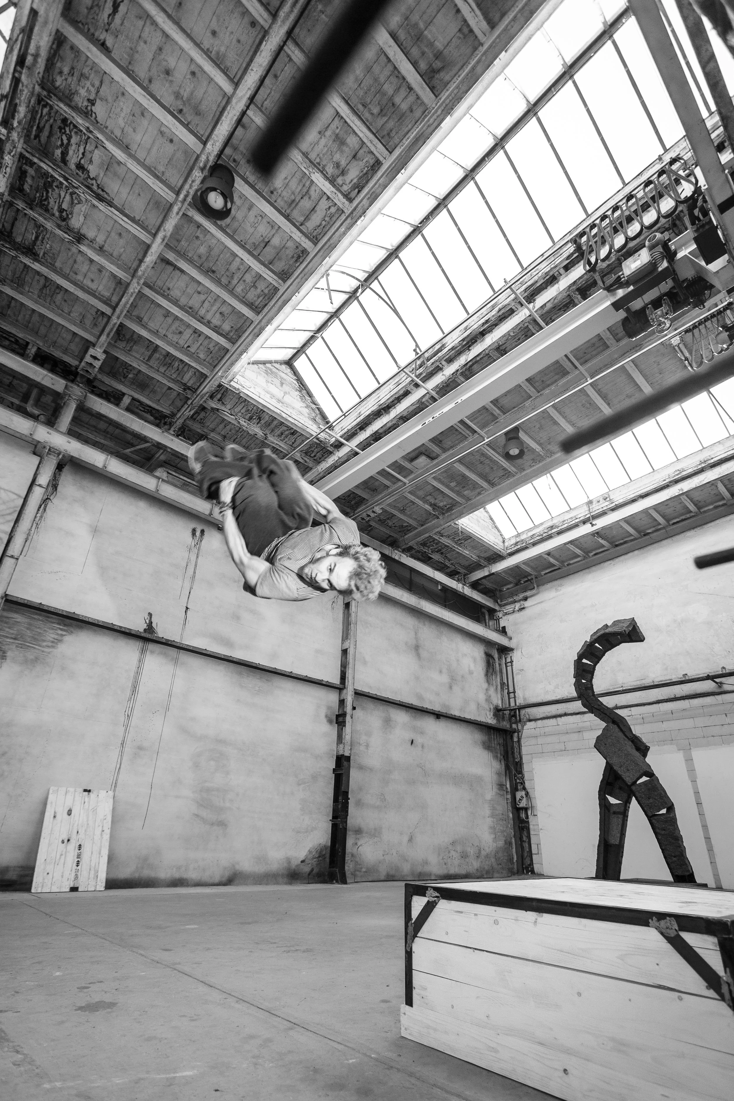 Mann, der im Sprung in einer Industriehalle springt, mit Skulptur und Kisten im Vordergrund, Decken mit großen Fenstern im Hintergrund, schwarz-weiß Fotografie.