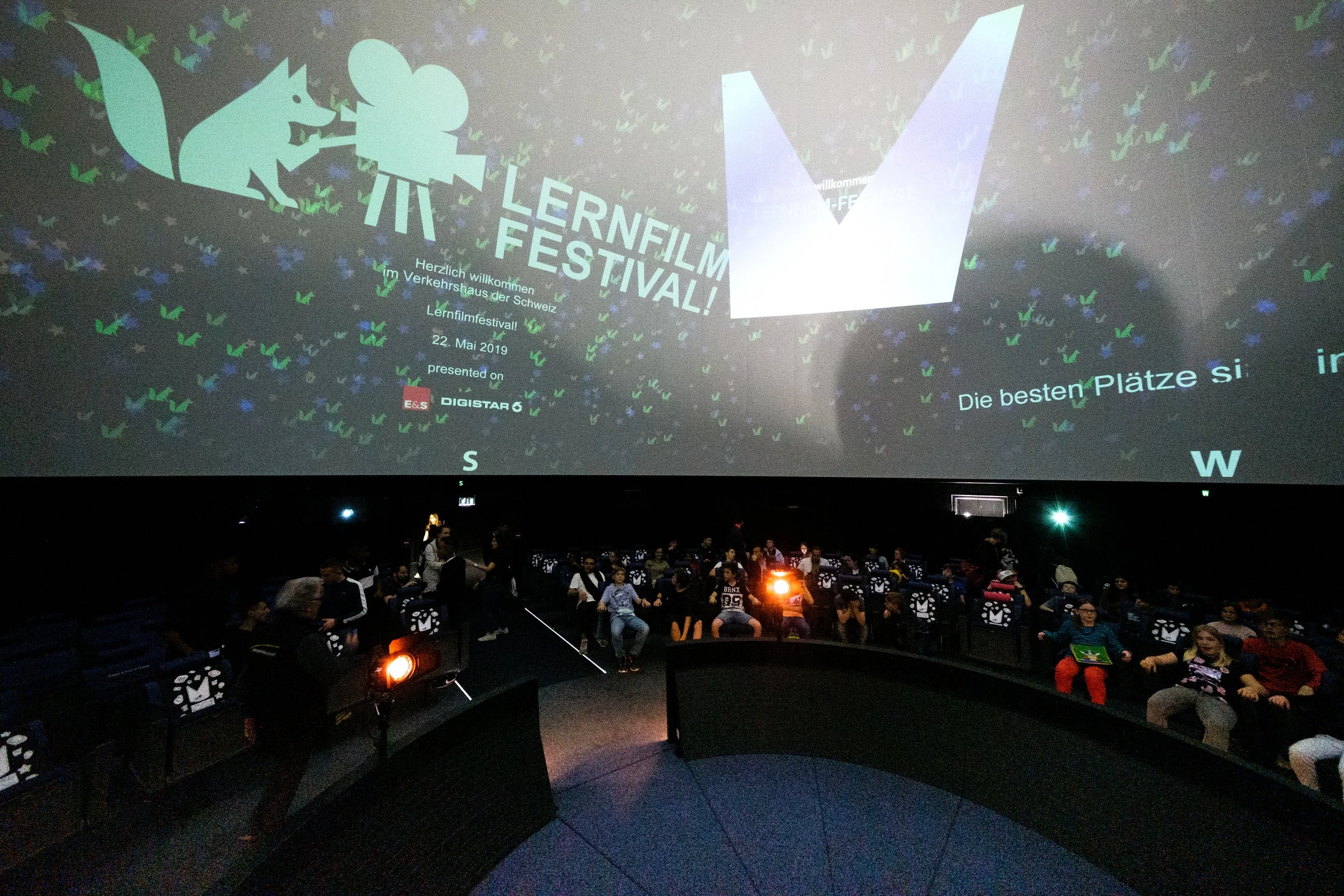 Innenaufnahme eines Kinos mit einem großen Bild groß an der Wand, das das Lernfilmfestival Schwarzwald bewirbt. Es gibt viele Sitzplätze und einige Menschen sitzen in den Kinosesseln. Bei den Leuten sind zwei den Film schauende Kinder, außerdem stehe