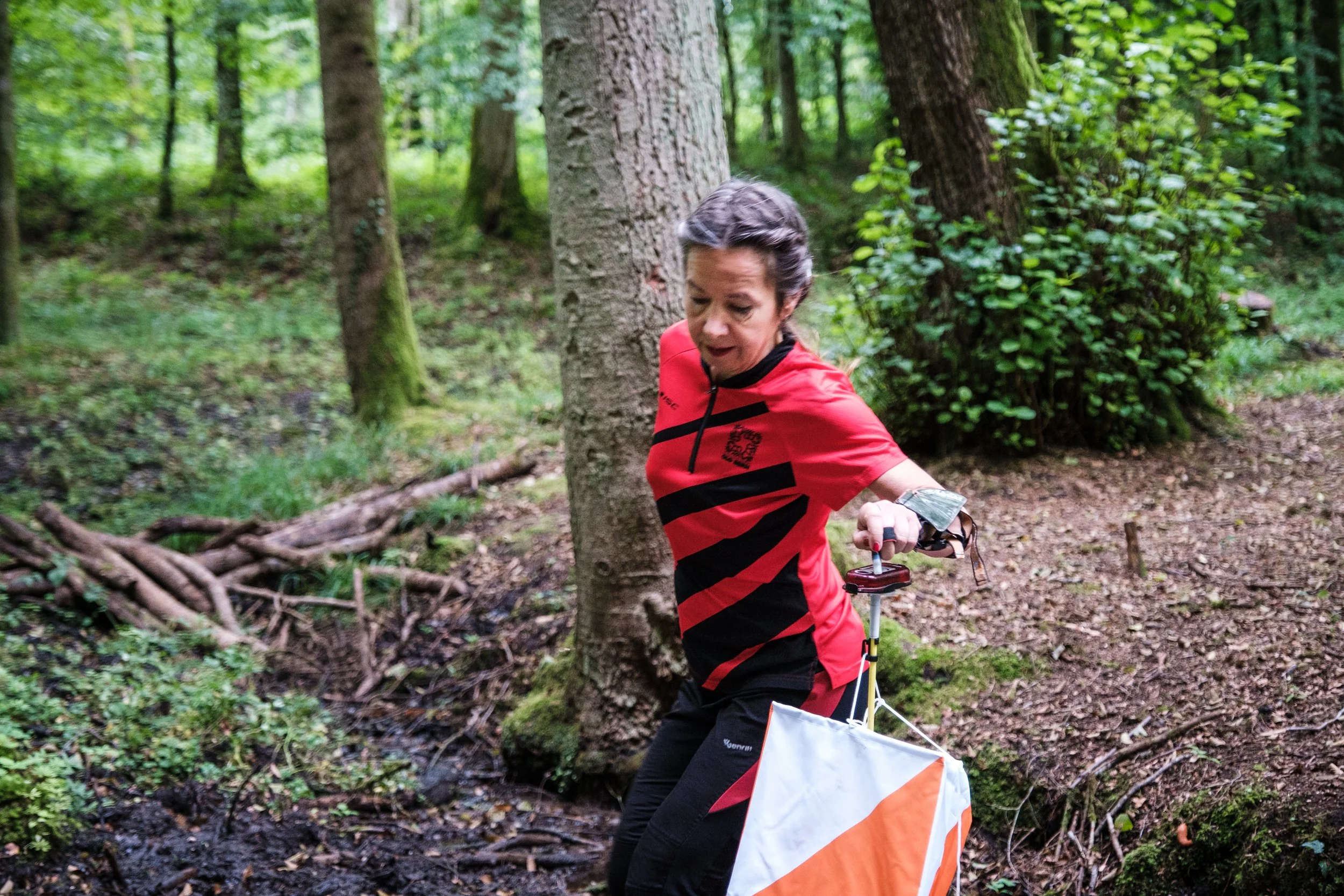 Frau in rotem und schwarzen Sportoutfit beim Flaggeneinsatz im Wald, vermutlich beim Orientierungslauf.
