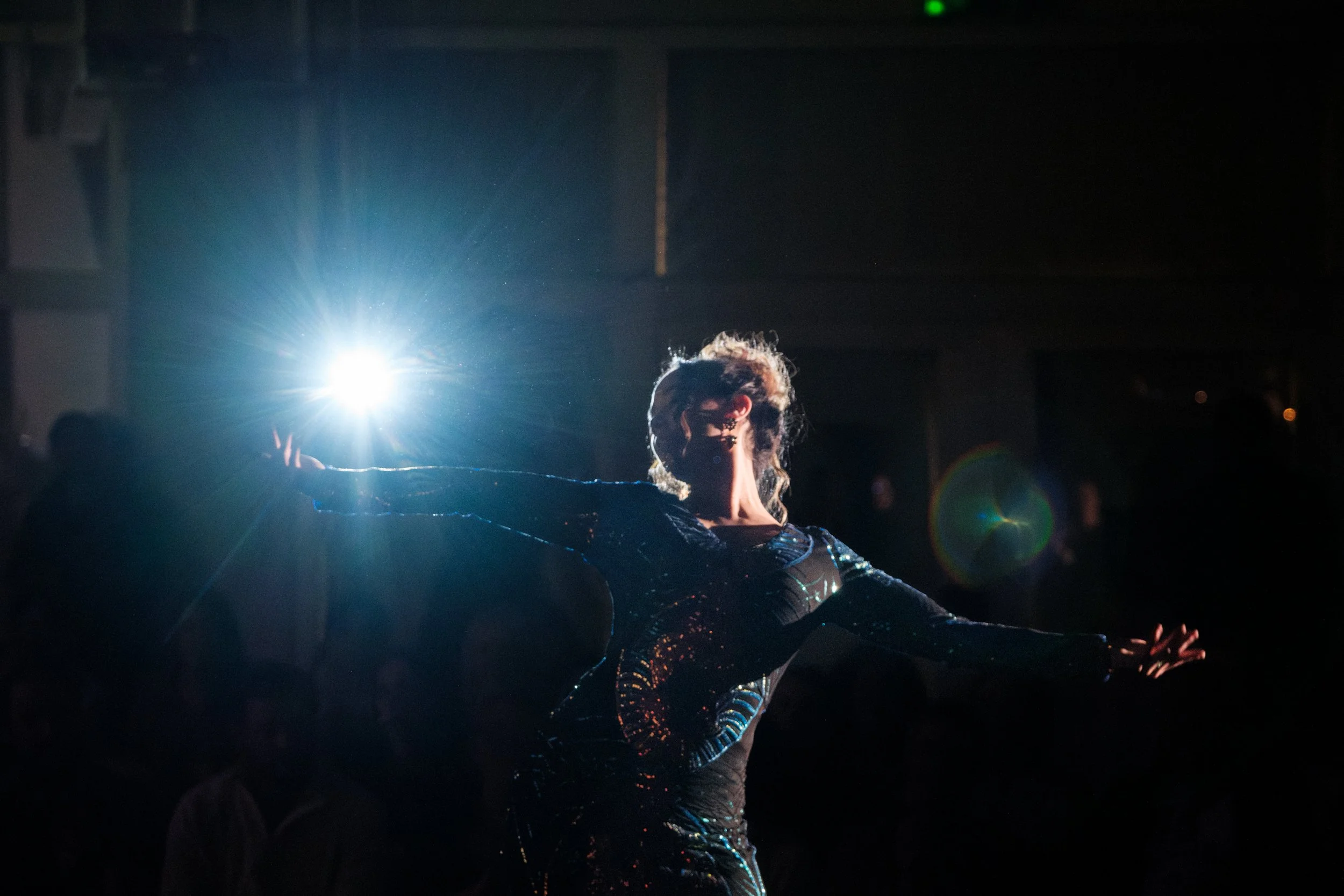 Tänzerin in einem schwarzen, glitzernden Kleid führt eine Performance auf der Bühne aus, während eine helle Lichtquelle im Hintergrund leuchtet.