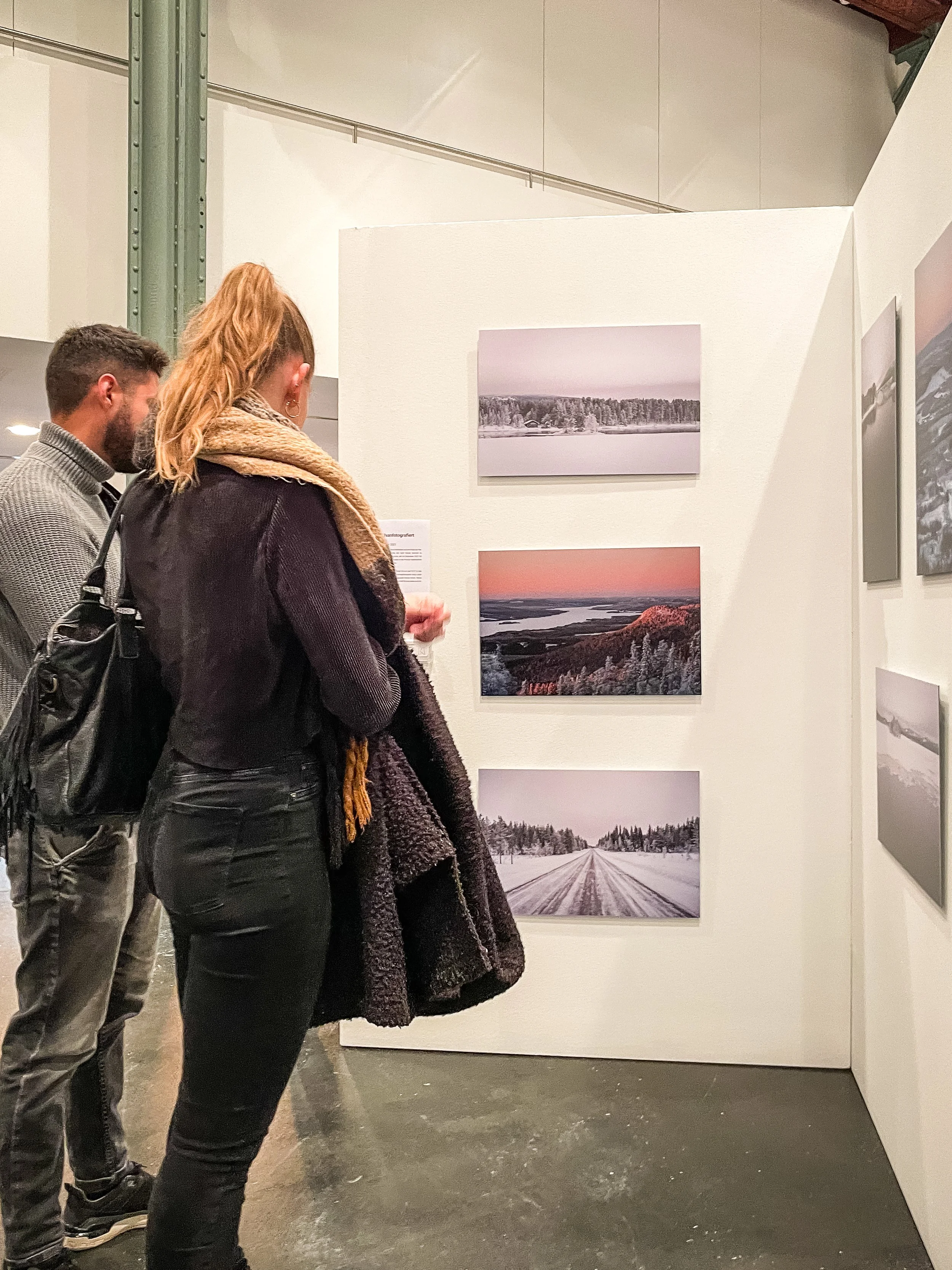 Zwei Menschen betrachten Landschaftsfotografien in einer Galerie, die Bilder zeigen Schnee, Wasser und Berge in winterlichen Landschaften.